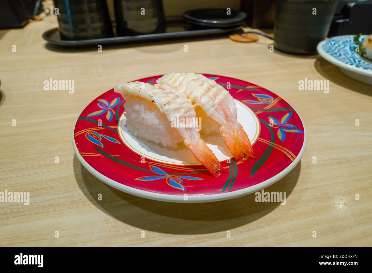 Due pezzi di sushi nigiri di gamberi sono serviti su un piatto rosso vivace con motivi floreali blu e oro, adagiati su un bancone di legno chiaro in un moderno ristorante di sushi Osaka. Foto Stock