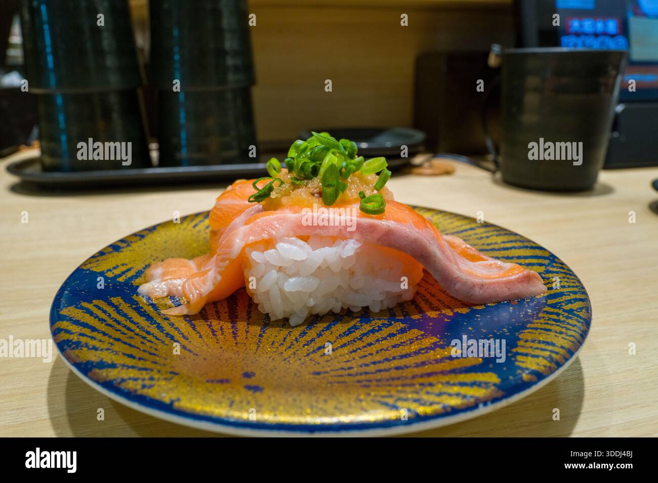 Una fetta di salmone fresco drappeggiata sul riso aceto è condita con cipolla verde tritata e salsa, servita su un vivace piatto blu e dorato in un moderno ristorante sushi di Osaka. Foto Stock
