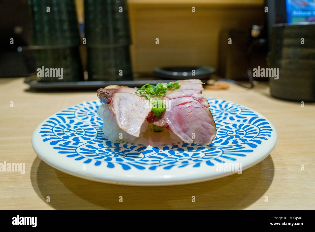 Un pezzo di sushi di orata di mare condito con cipolla verde a fette poggia su un piatto bianco caratterizzato da audaci motivi floreali blu, adagiato su un bancone di legno chiaro in un moderno ristorante sushi di Osaka. Il pesce fresco e la colorazione vivace creano una presentazione visivamente attraente e appetitosa. Foto Stock