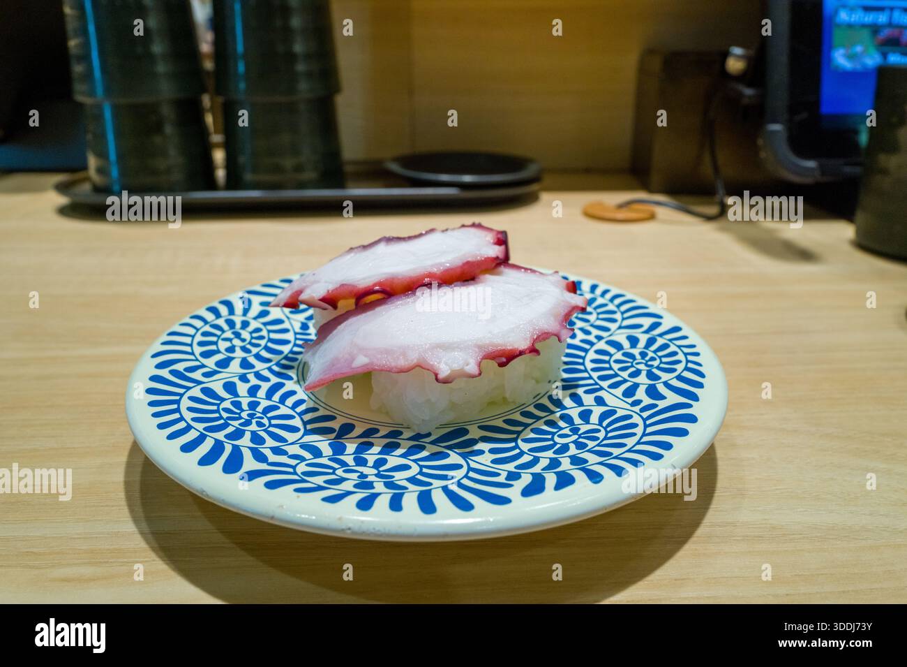 Due pezzi di sushi di polpo nigiri sono disposti su un piatto bianco con audaci motivi circolari blu, adagiati in cima ad un bancone di legno chiaro in un ristorante sushi contemporaneo di Osaka. L'immagine cattura le consistenze di pesce fresco e un'atmosfera da pranzo pulita e invitante. Foto Stock