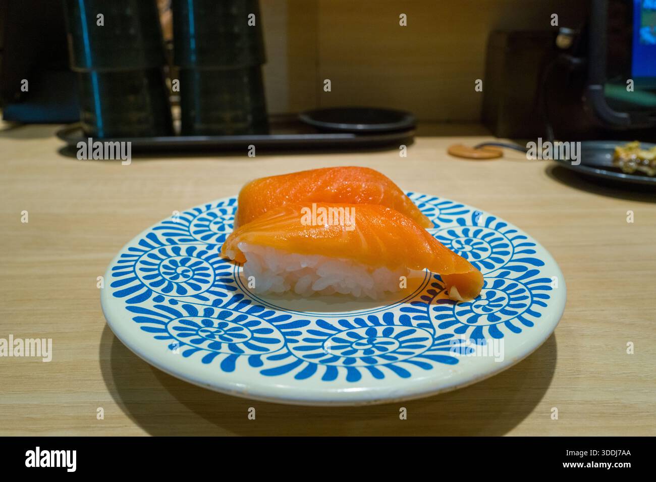 Due pezzi di sushi nigiri di salmone fresco poggiano su un piatto bianco con audaci motivi floreali blu, adagiati su un bancone di legno chiaro in un moderno ristorante sushi di Osaka. La scena evidenzia colori vivaci e una presentazione minimalista. Foto Stock