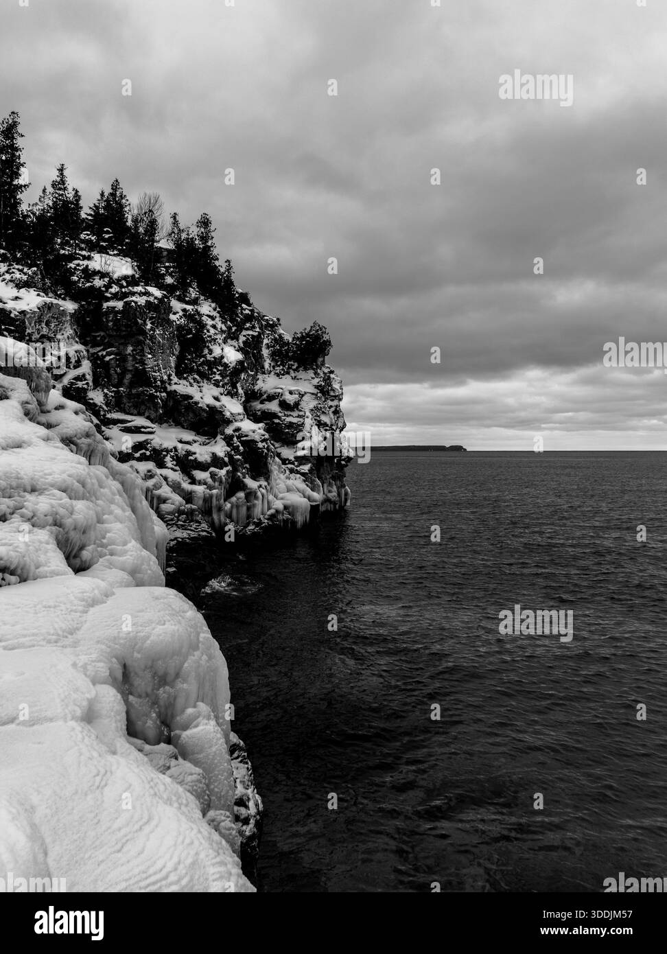 Costa invernale bianca e nera con rocce innevate e acque scure sotto pesanti nuvole. Foto Stock