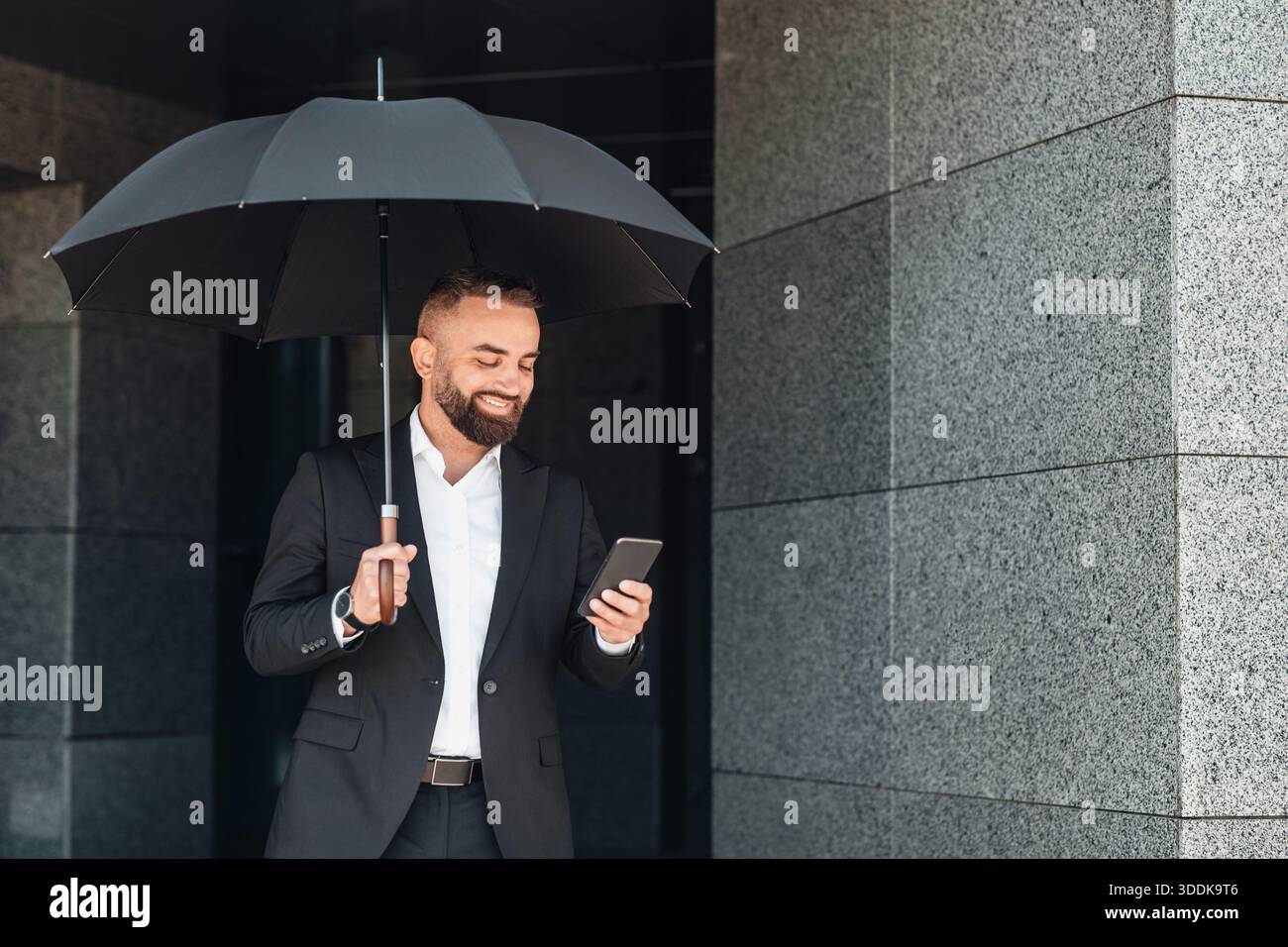 Uomo d'affari felice e maturo che usa lo smartphone tenendo in mano l'ombrello nero durante la passeggiata in città il giorno della pioggia Foto Stock