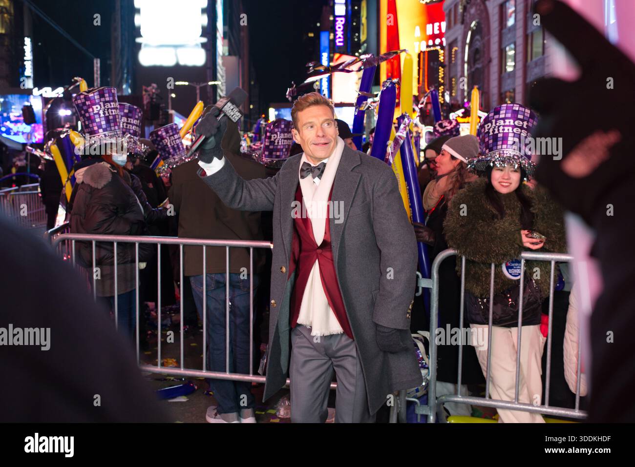 Un uomo con un cappotto grigio, un papillon e un gilet Bordeaux tiene un microfono e le onde in un evento notturno in una città affollata. Foto Stock