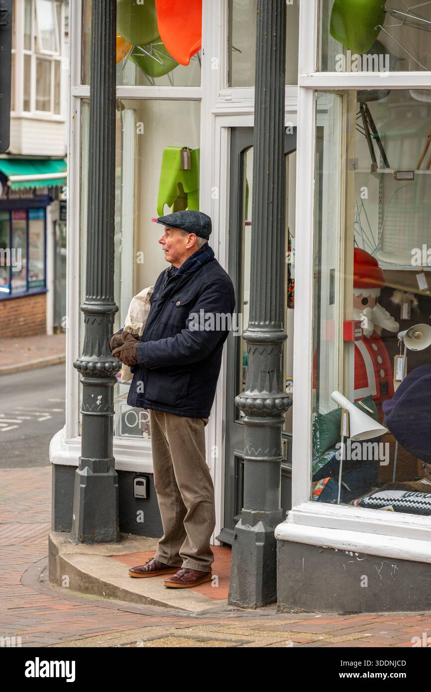 Uomo anziano o anziano che indossa abiti invernali e un berretto piatto da solo all'ingresso di un negozio vintage a Ventnor, sull'isola di wight, Regno Unito. Foto Stock