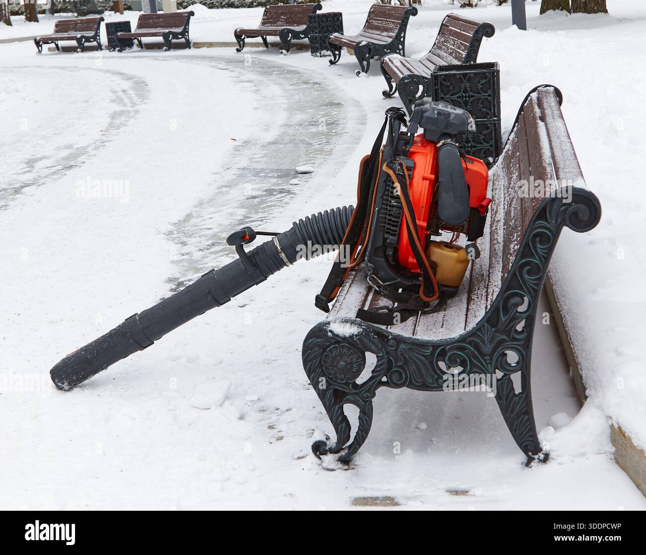 Soffiatore di foglie collocato su una panchina innevata in un parco, che mette in evidenza le attrezzature per la manutenzione invernale con un tortuoso sentiero innevato nelle vicinanze Foto Stock