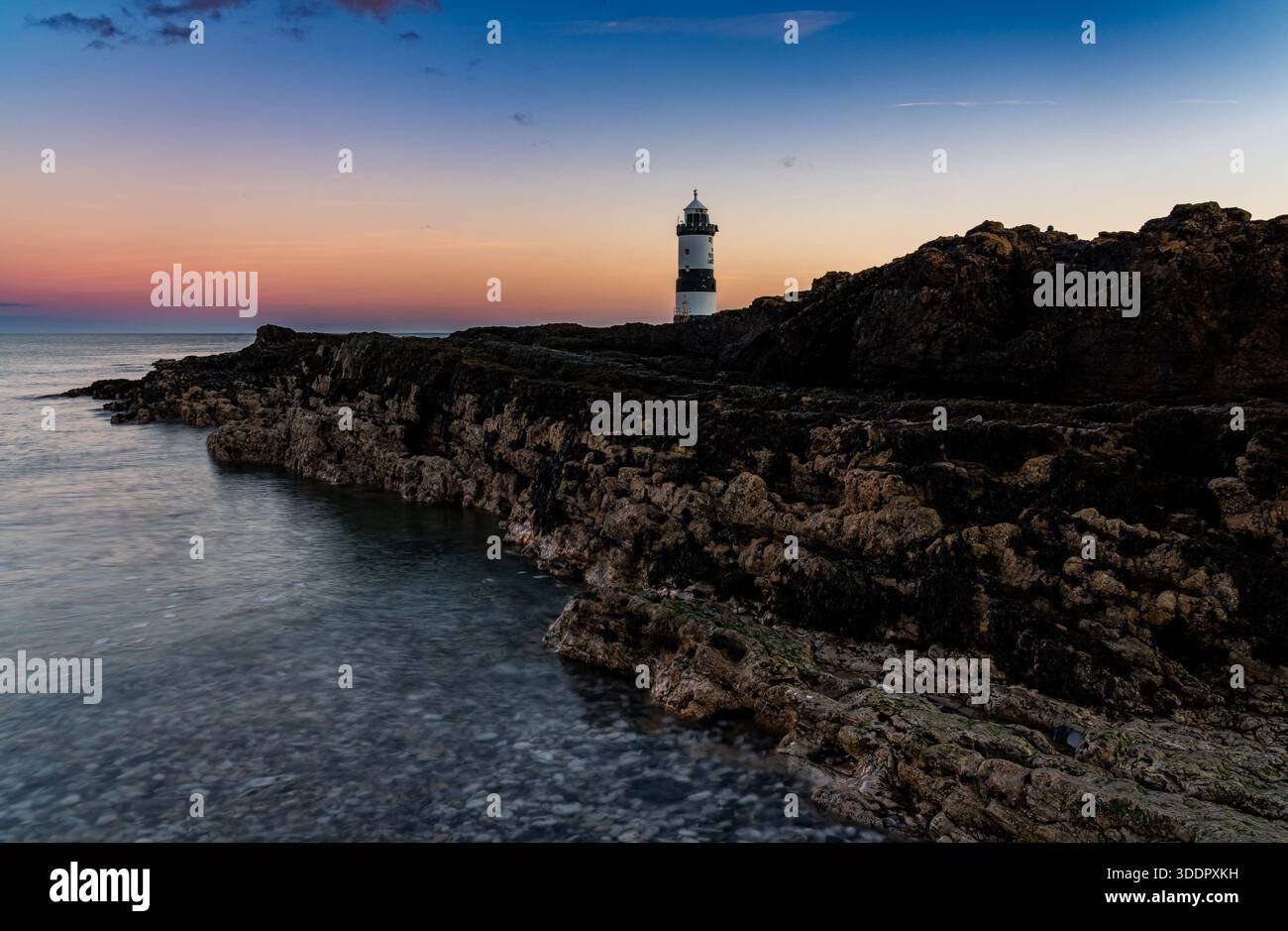 Faro di Penmon Point all'alba sopra una costa rocciosa e calme acque costiere Foto Stock