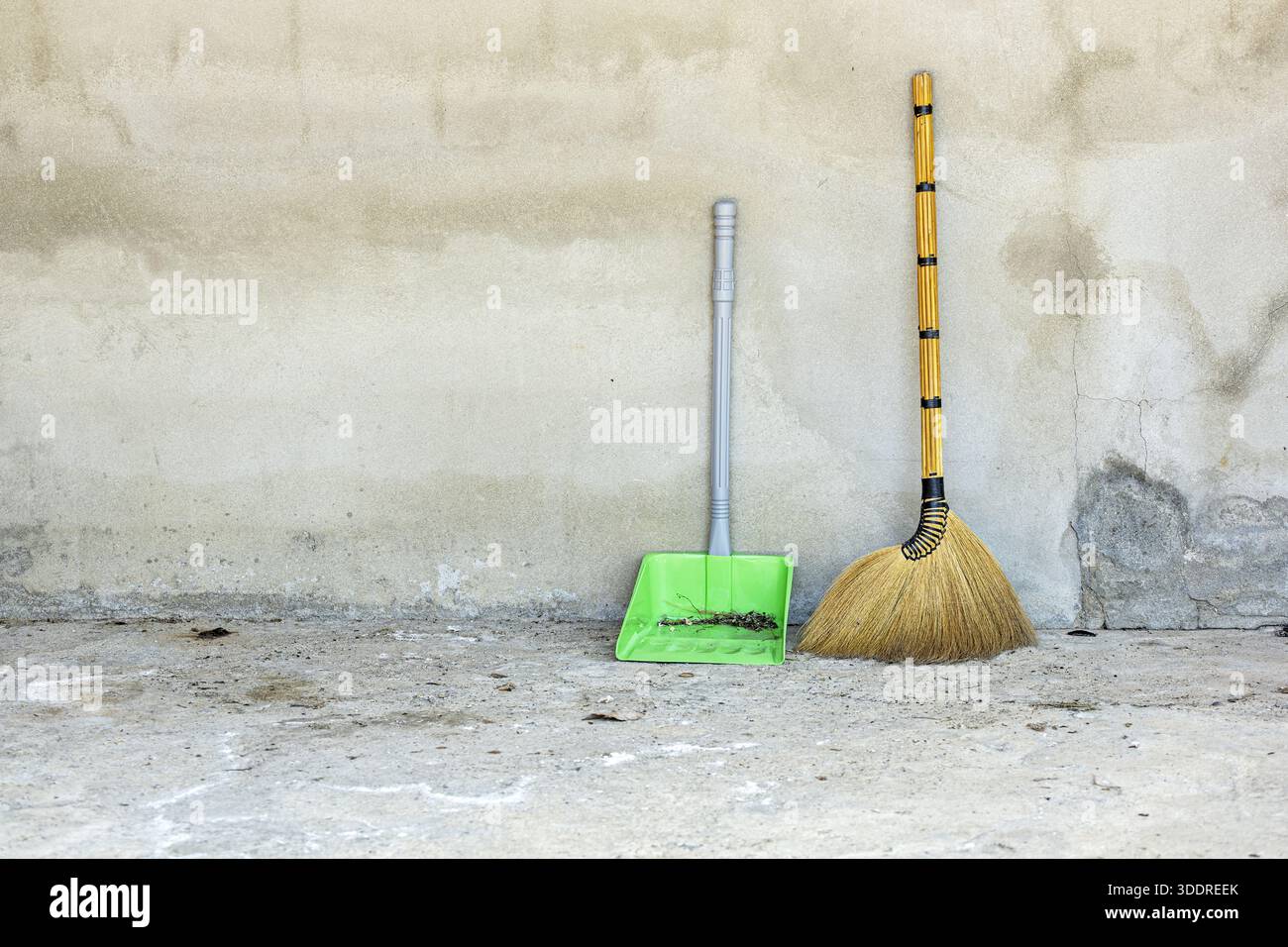 Una scopa tradizionale in erba e una paletta in plastica verde riempita di detriti si appoggiano a una parete in cemento armato resistente agli agenti atmosferici e al pavimento sporco. La scena Foto Stock