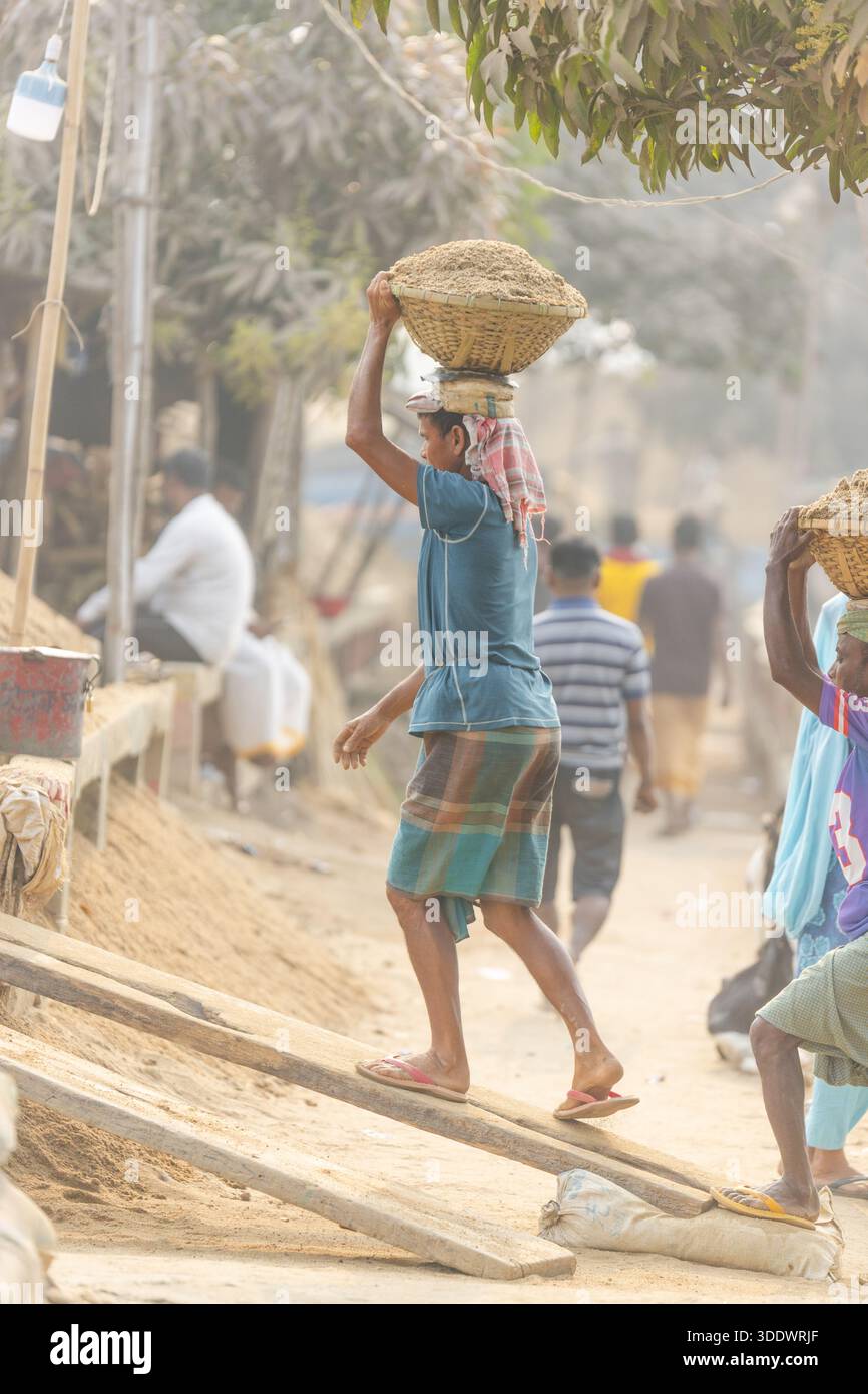 Dhaka, Bangladesh - 19 febbraio 2025: Vista di un operaio edile che bilancia un cesto di sabbia sulla testa, camminando lungo un sentiero polveroso sotto il sole nebbioso Foto Stock