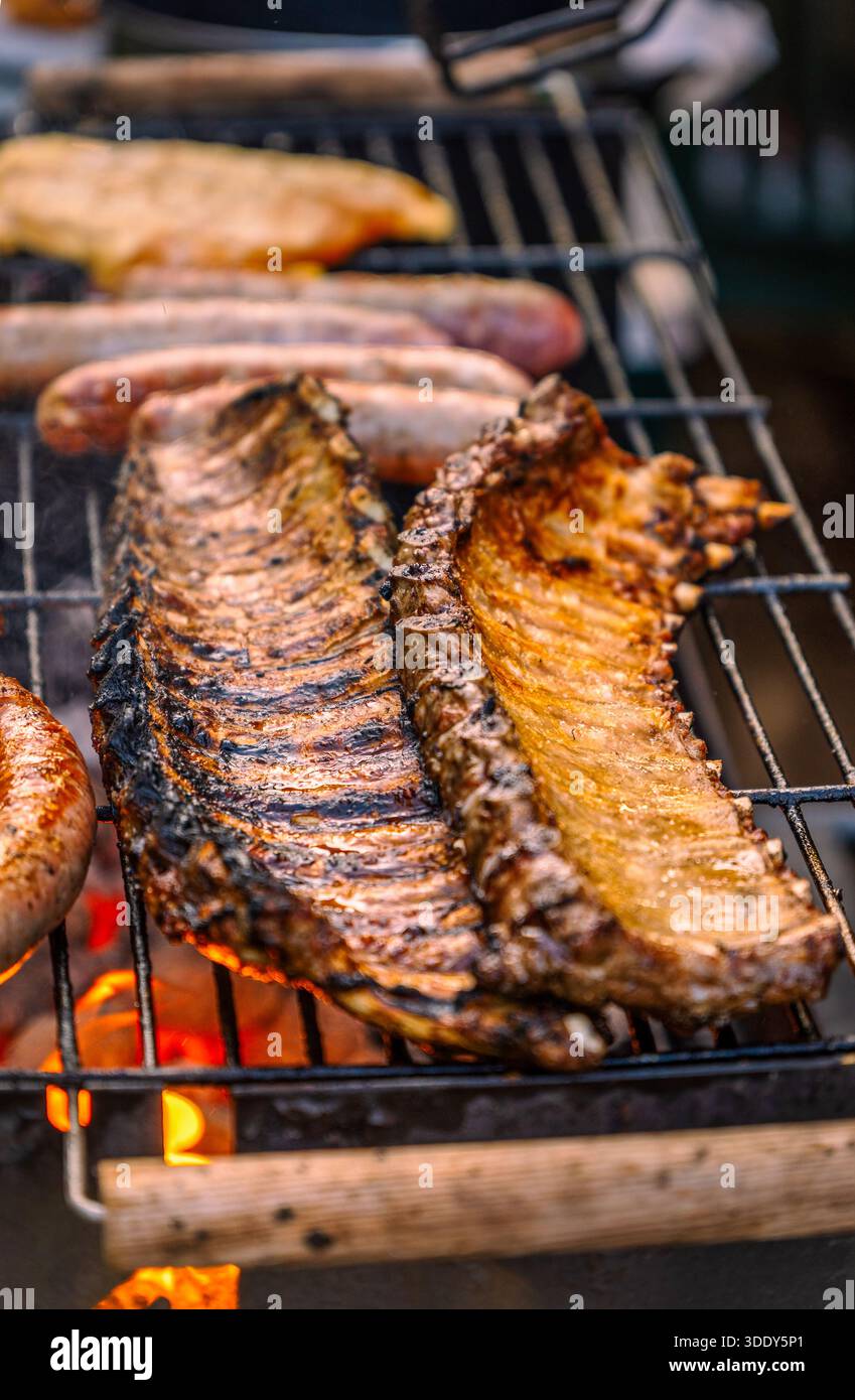 Primo piano verticale di due costolette di maiale dorate che arrostono lentamente su una griglia per barbecue con salsicce e filetti di pollo sullo sfondo. Foto Stock