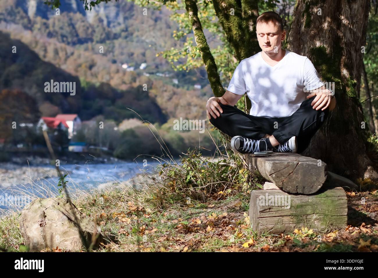 Un maschio adulto si rilassa a occhi chiusi e palme giù sulle gambe mentre è seduto sul fiume nel paesaggio autunnale Foto Stock