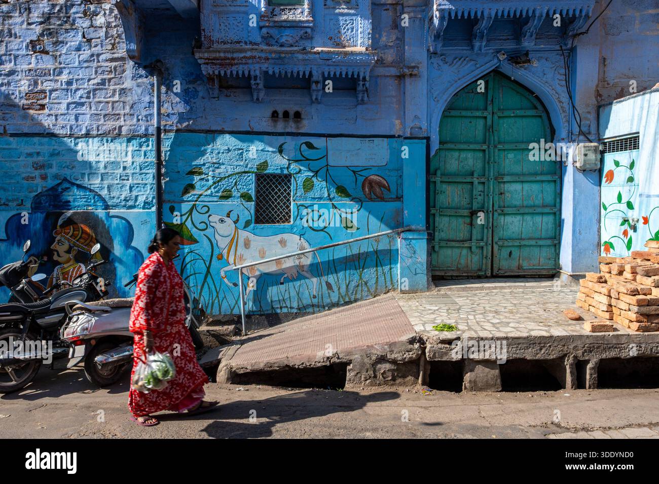 Jodhpur, India - 22 dicembre 2025: Una donna con un vestito colorato passa di fronte a un edificio abbandonato di colore azzurro, senza altre persone. Foto Stock