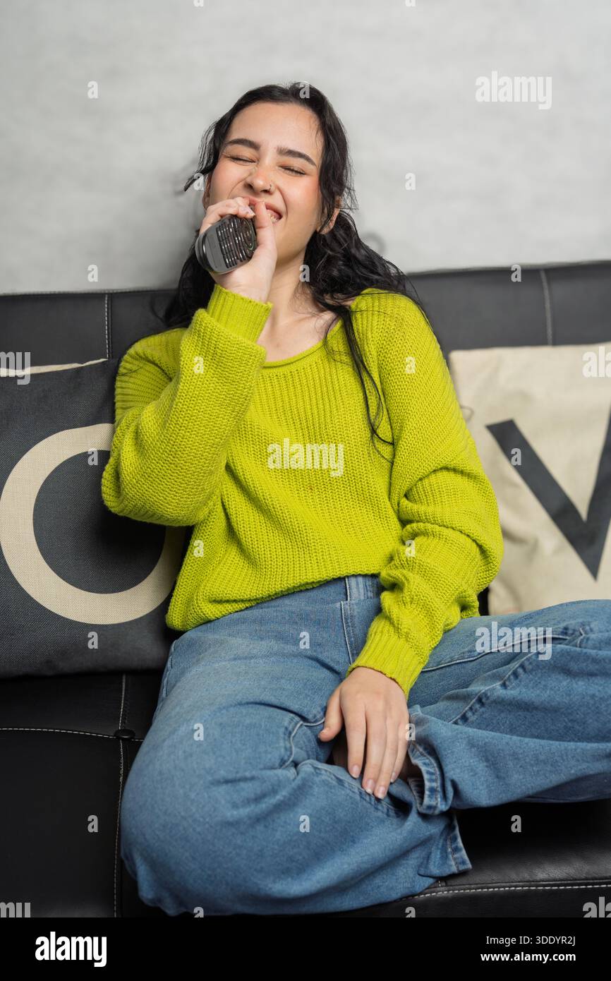 Giovane donna che canta felicemente in un telecomando e si diverte sul divano di casa Foto Stock