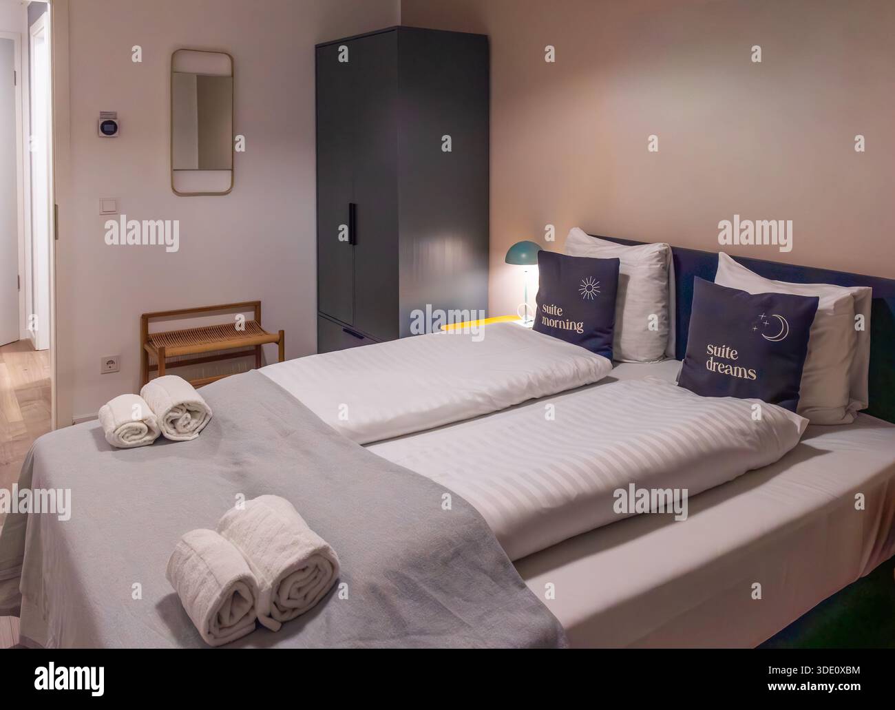 Immagine orizzontale grandangolare di una camera da letto minimalista ordinata con letto grigio, lenzuola bianche, asciugamani rotolati, armadio e illuminazione soffusa Foto Stock