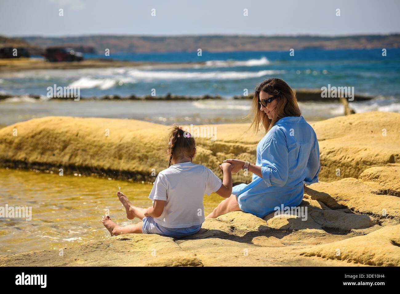 Una donna e una ragazza si siedono sulle rocce calcaree gialle in riva al mare, tenendosi per mano e spruzzando acqua con i piedi sotto il sole luminoso, vista sul retro. Foto Stock