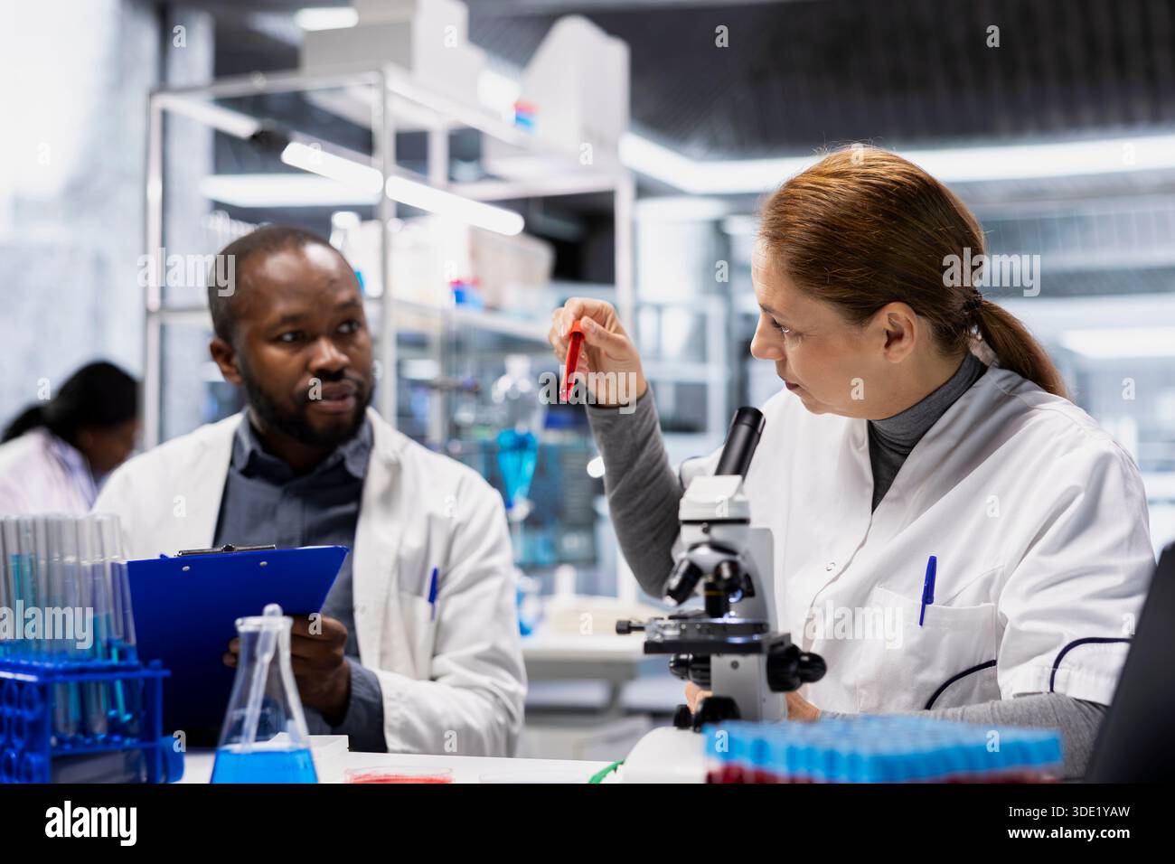 Ricercatrice donna e collega di laboratorio che analizza campioni di sangue per reazioni avverse da farmaci. Team di scienziati brainstorming, valutazione di flaconcini fluido sanguine per garantire gli standard di sicurezza dei farmaci, telecamera A. Foto Stock