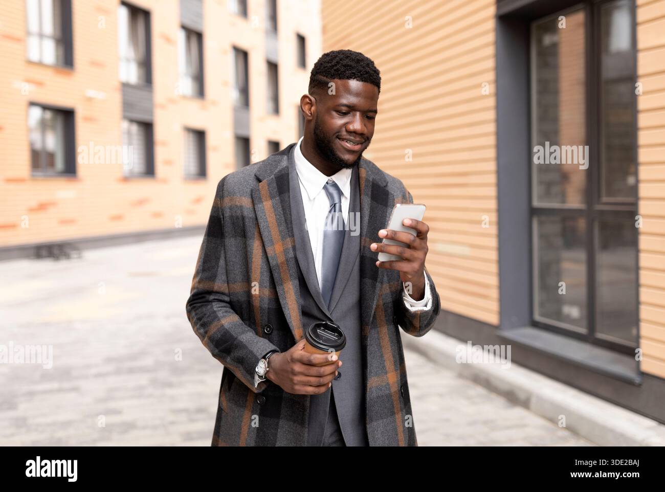 Felice uomo d'affari nero texting sul cellulare e bere caffè takeaway, a piedi fuori ufficio edificio Foto Stock