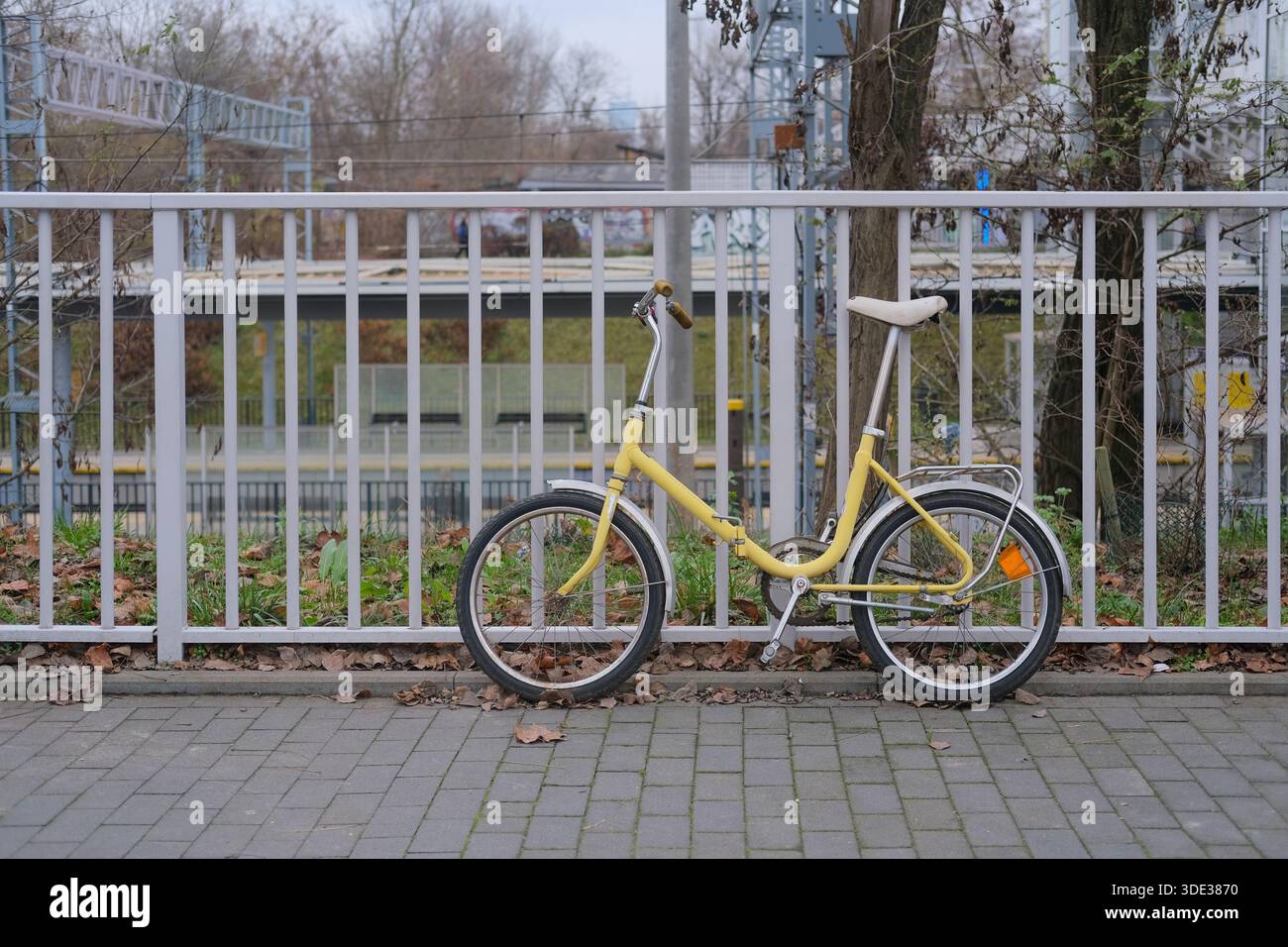 Vista laterale di una bicicletta passante gialla parcheggiata su un terreno pavimentato accanto a una ringhiera metallica bianca. Luce diurna, messa a fuoco profonda, outdoor urbano. Foto Stock