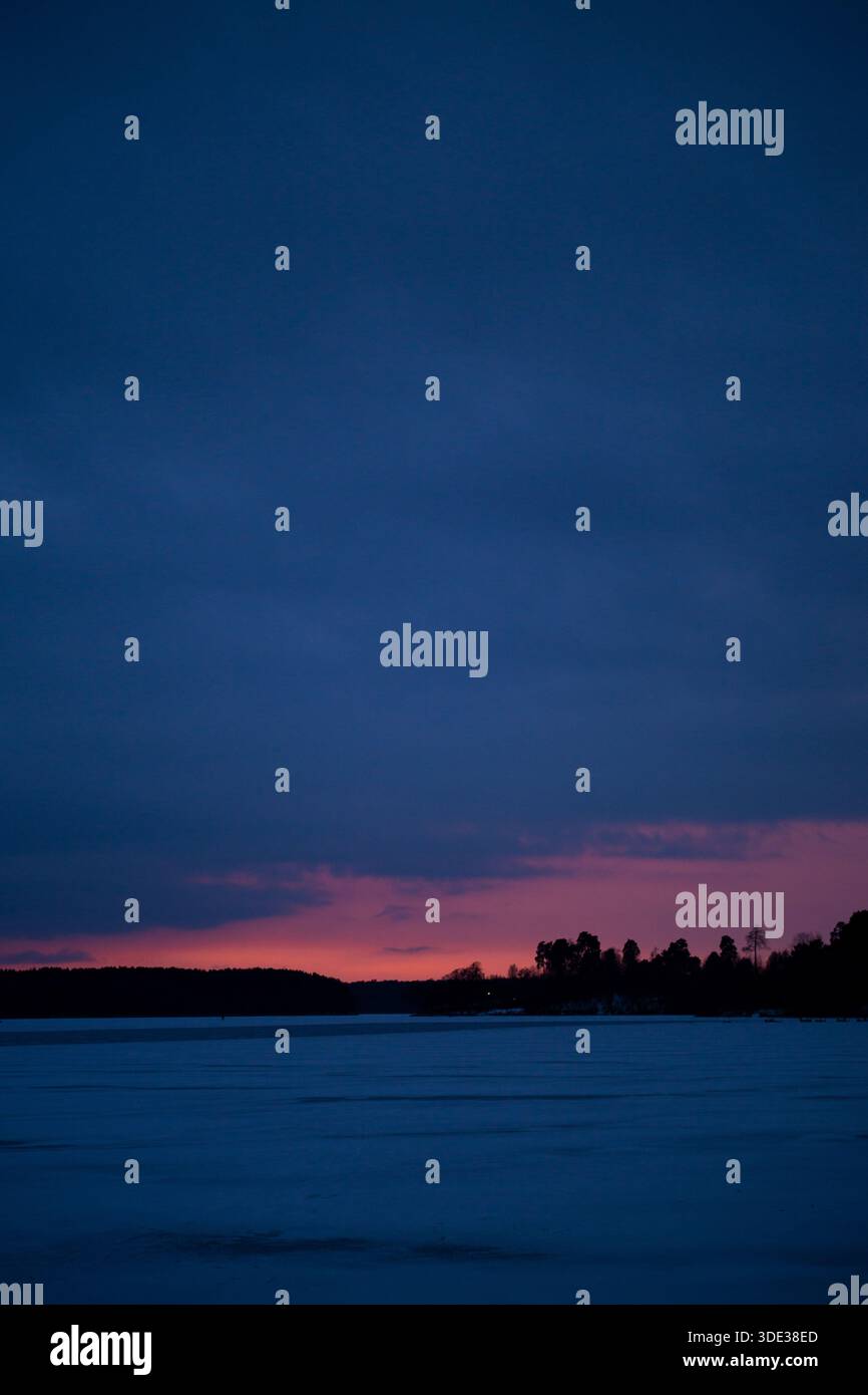 Bellissimo tramonto viola caldo in un parco vicino a un fiume ghiacciato, a una foresta lontana, a nuvole blu e neve Foto Stock
