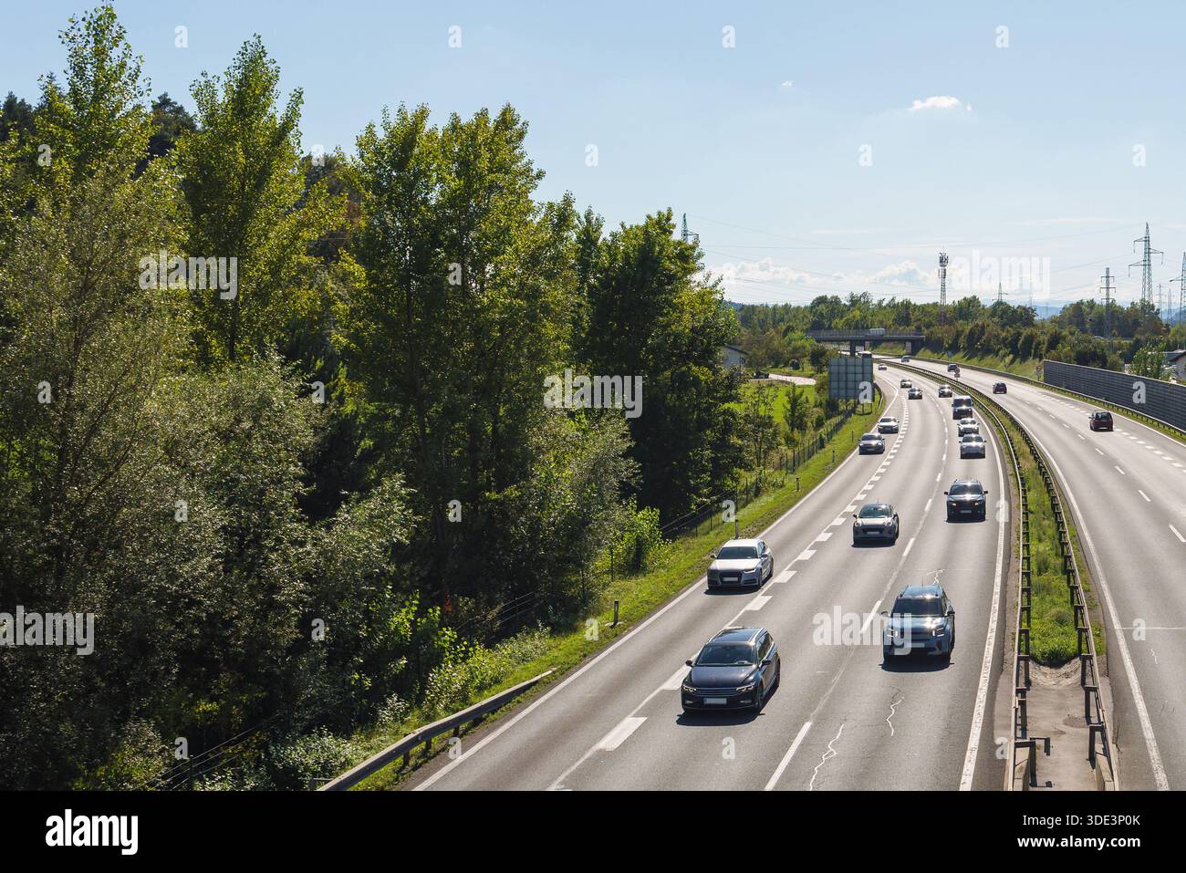 Le auto si spostano lungo un'autostrada trafficata circondata da alberi e infrastrutture moderne. Questa scena mostra gli spostamenti quotidiani e la vita urbana. Foto Stock