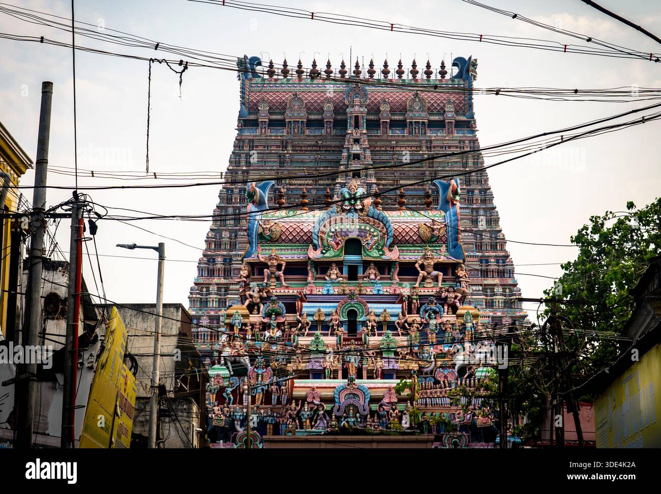 Colorato gopuram del tempio indù con intricate sculture e incisioni, circondato da fili ed edifici urbani, sotto un cielo nuvoloso. Foto Stock