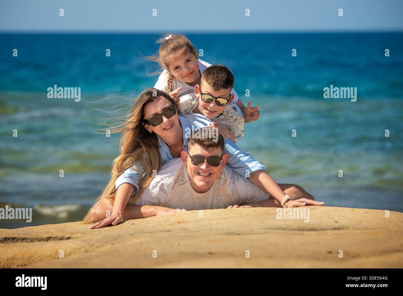 Una famiglia gioiosa di quattro persone si diverte a costruire una piramide umana sulle rocce costiere di arenaria sullo sfondo del mare blu. Foto Stock