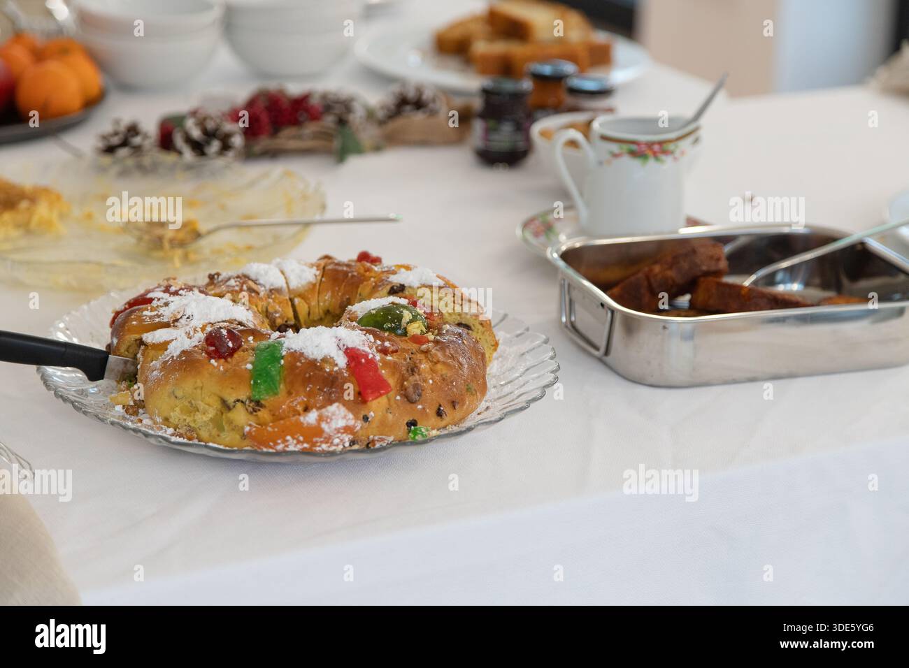 Un tavolo bianco è allestito con tradizionali dolci natalizi, con una torta a fette con frutta candita, toast alla francese, mandarini freschi e mele Foto Stock