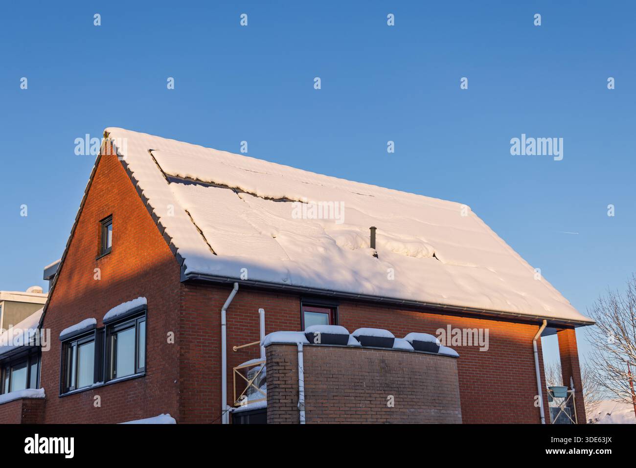 Tetto di una casa in mattoni rossi nei Paesi Bassi con pannelli solari parzialmente coperti da uno spesso strato di neve durante una soleggiata giornata invernale Foto Stock