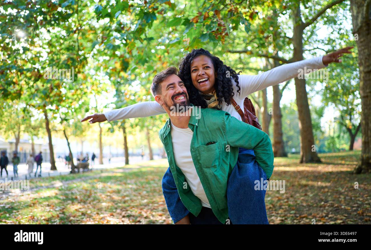 Coppia giovane felice e variegata che si diverte con un giro in piggyback nel parco. Foto Stock