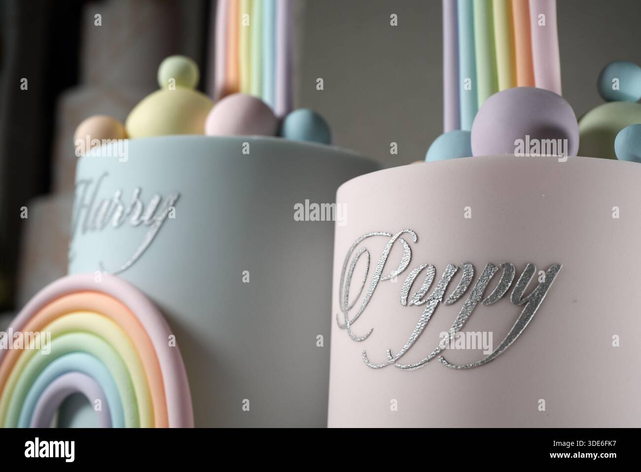 Un'esibizione di torte di compleanno con due livelli separati, i nomi delle scritte in argento e un alto ponte arcobaleno pastello, presentato su un piedistallo bianco Foto Stock