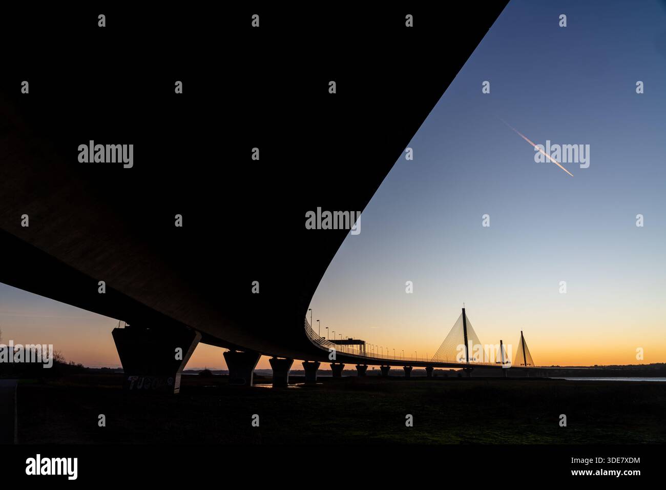 Vista sotto il Mersey Gateway Bridge all'alba, con l'ampio sottoscocca che conduce verso le campate strallate e un aereo jet che lascia un cont Foto Stock