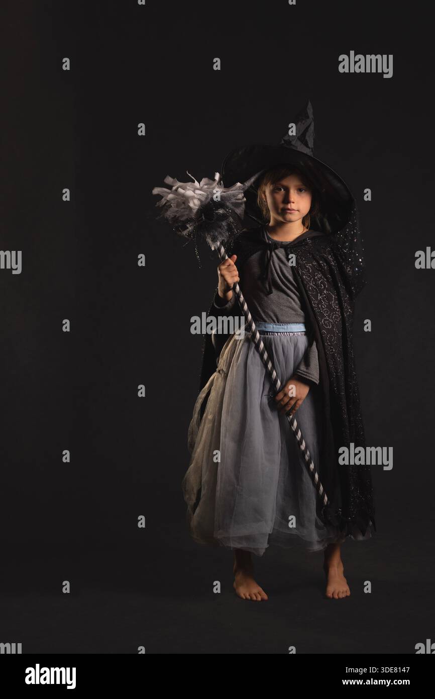 Bambino vestito da strega che tiene in mano il personale decorativo in uno studio scuro, indossa cappello, mantello e gonna in tulle per halloween o carnevale Foto Stock