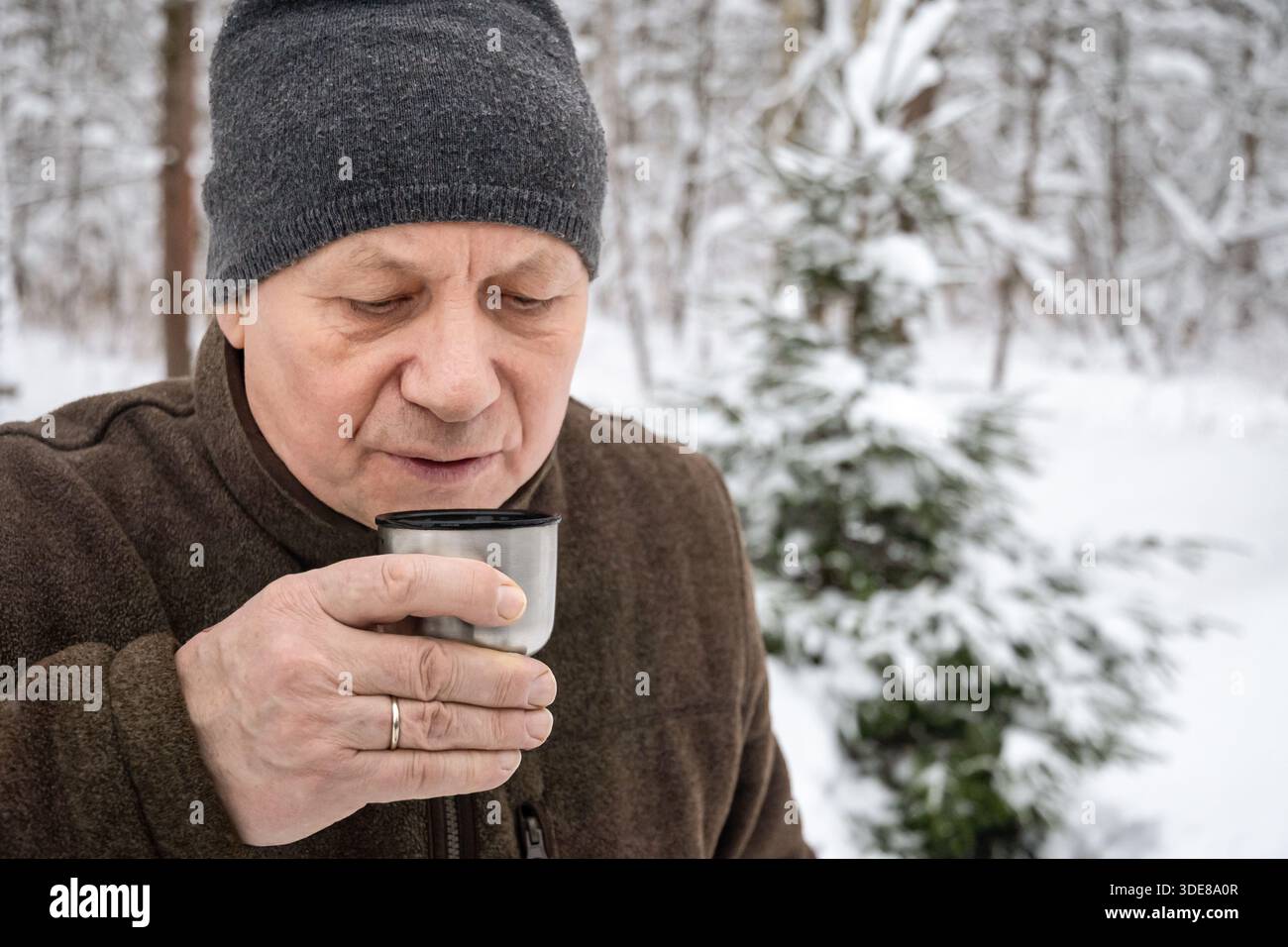 Uomo anziano che tiene una tazza di metallo da campo e beve bevande calde nella foresta invernale innevata. Momenti all'aperto accoglienti, relax e calore nelle giornate fredde. Foto Stock