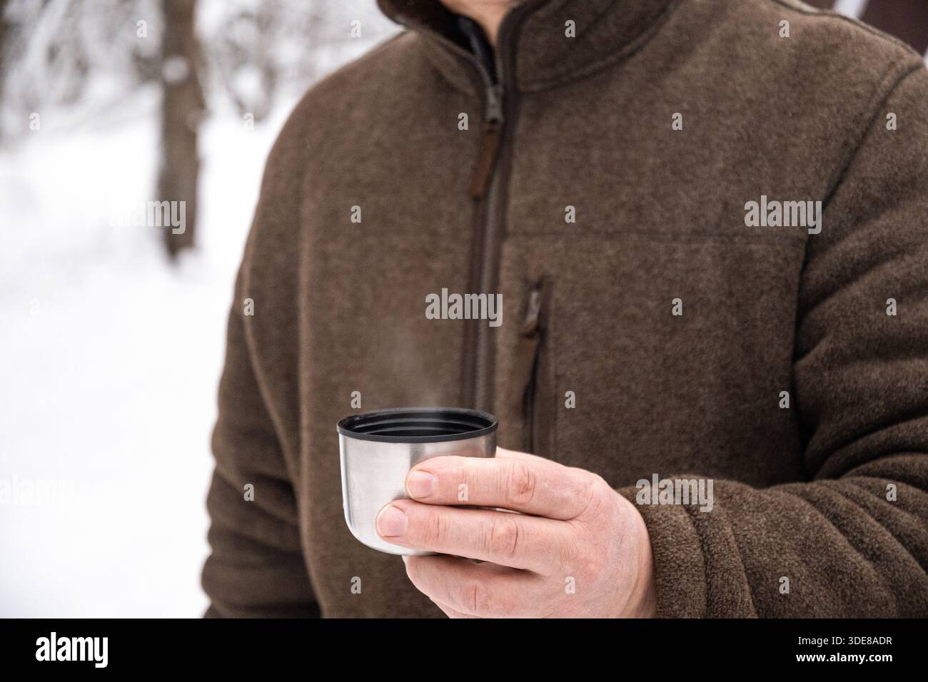 Primo piano di un uomo che tiene in mano una tazza di acciaio inossidabile con caffè caldo o una bevanda mentre si trova all'aperto nella foresta invernale innevata. Bevanda calda a freddo Foto Stock