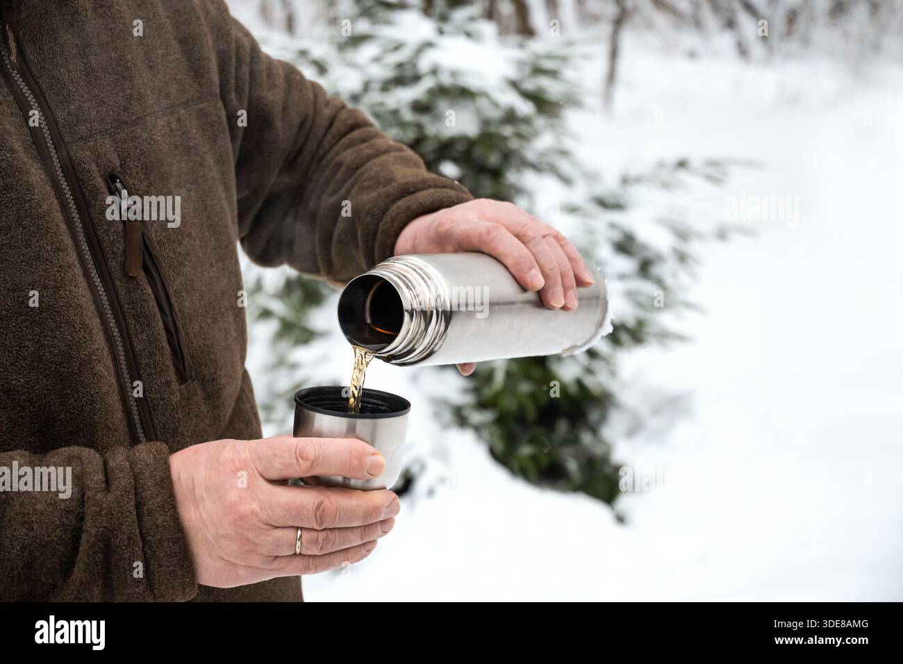 Mani di un uomo anziano che versa bevande calde da thermos in acciaio inox in una tazza di metallo all'aperto nella foresta invernale innevata. Bevanda calda in condizioni di freddo. Foto Stock