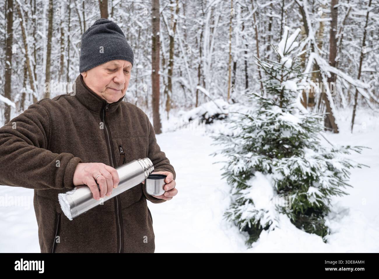 Uomo anziano in abiti caldi che versa tè o caffè caldo da una tazza thermos in acciaio inox mentre si trova in una foresta invernale innevata. Riposo all'aperto durante il freddo Foto Stock