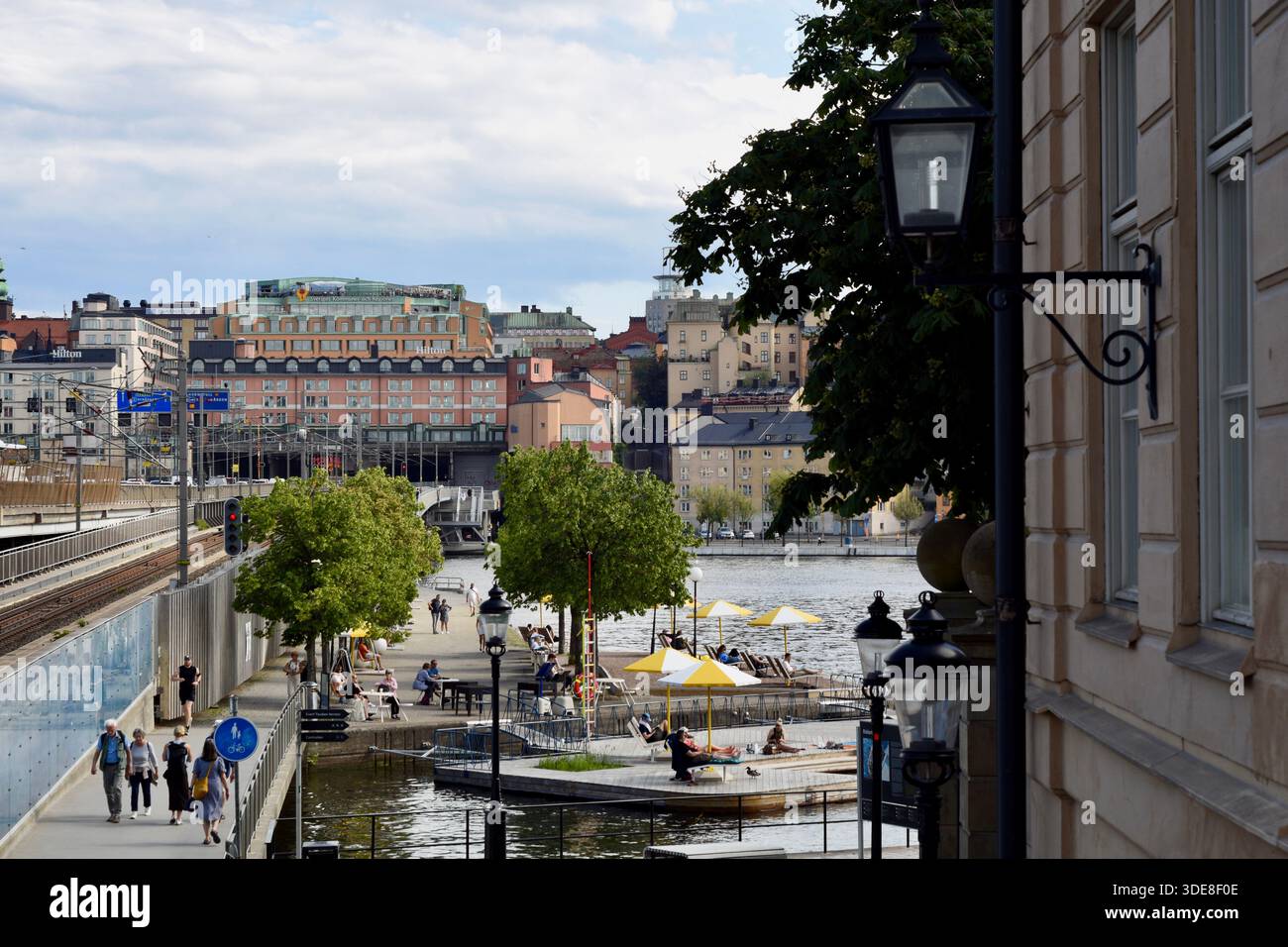 Persone che trascorrono del tempo lungo una passeggiata sul lungomare in una città durante l'estate, con pedoni, posti a sedere all'aperto ed edifici urbani sullo sfondo. Foto Stock