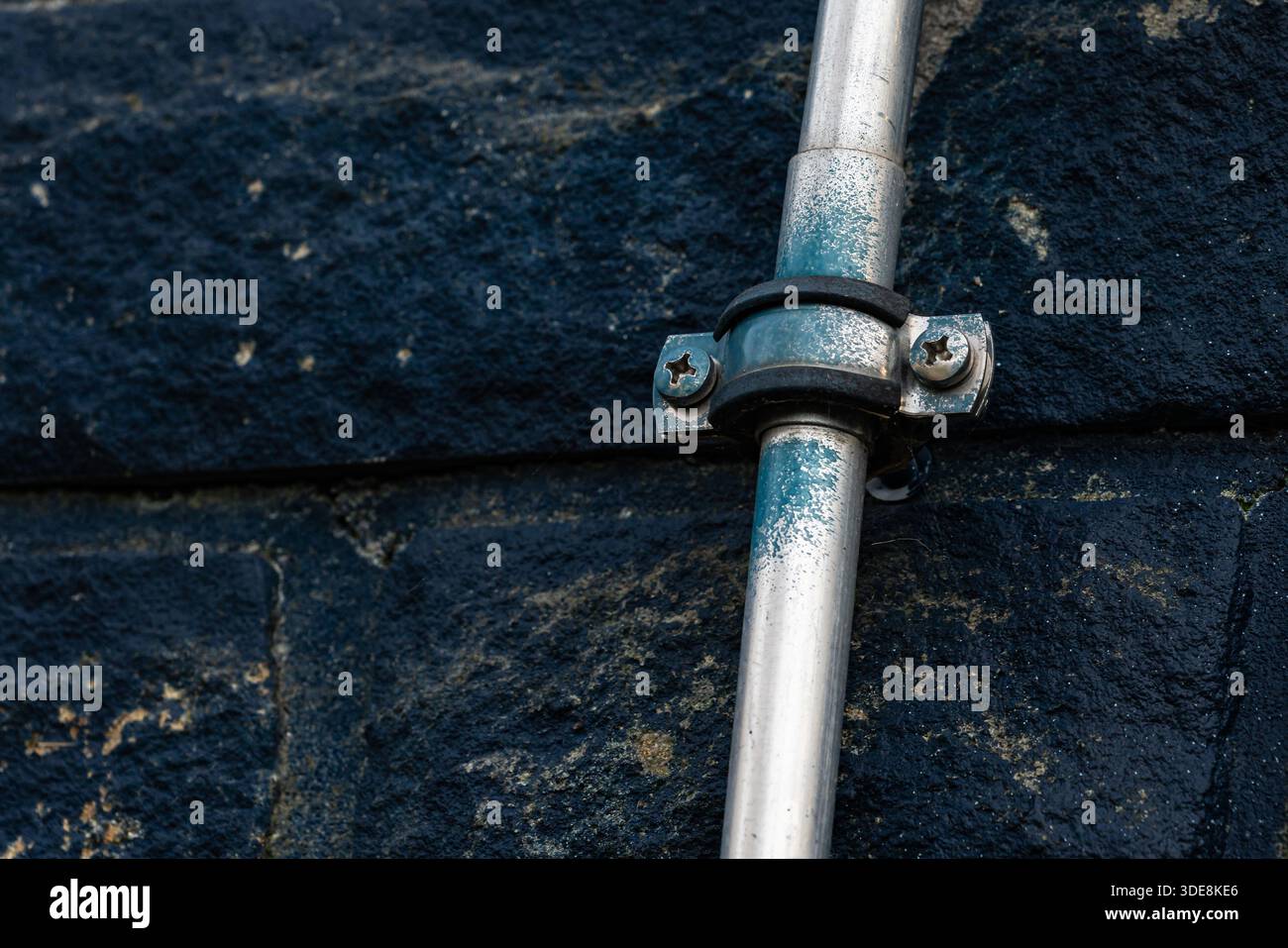 Un tubo metallico è fissato a un muro di pietra scura. Il tubo ha una finitura lucida ed è tenuto in posizione con morsetti e viti in metallo. Questa scena è comune Foto Stock
