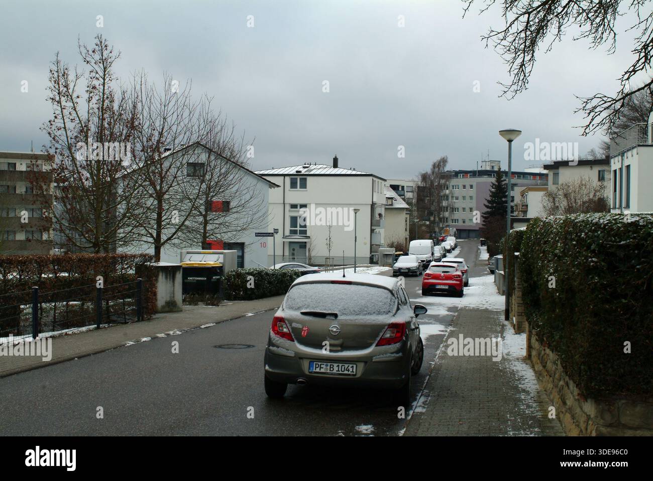 Strada residenziale innevata con auto, corsia in pendenza fiancheggiata da siepi e case, veicoli compatti parcheggiati lungo il marciapiede, cielo pallido coperto, Keukenhof Foto Stock