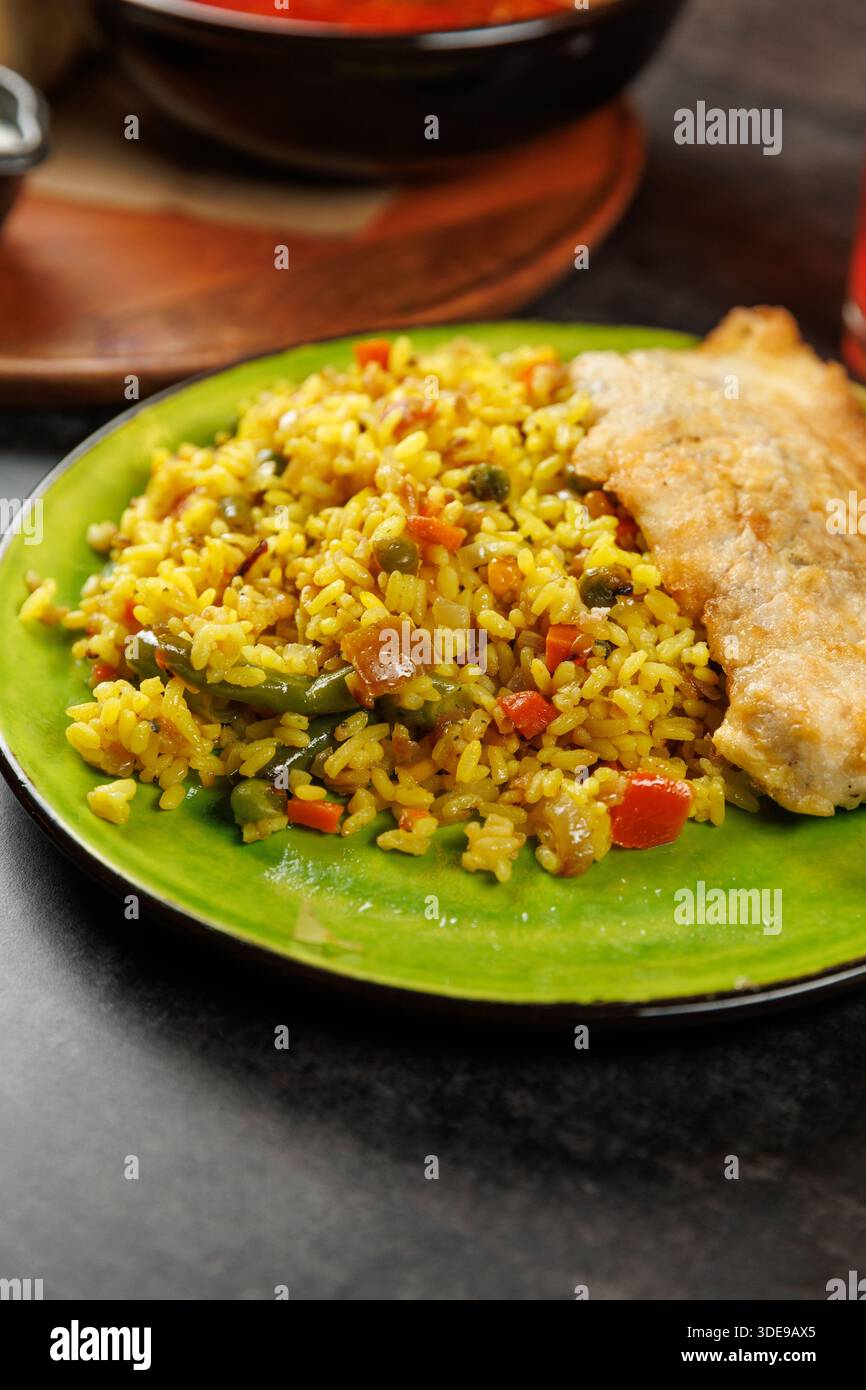 I piatti di riso mescolati con verdure e un pezzo di pesce fritto sono disposti su un piatto verde brillante. La colorata presentazione mostra la fusione di Foto Stock