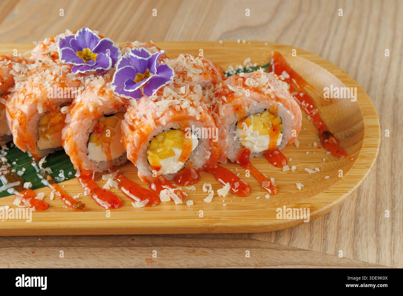 Gli involtini di sushi sono disposti ordinatamente su un piatto di legno. Gli involtini sono ripieni di salmone, uova, ricoperti di salsa e guarniti con fiori commestibili. PL Foto Stock