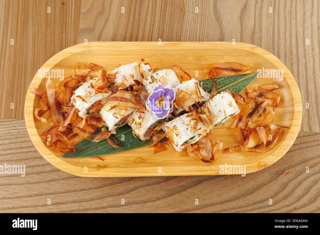 Un piatto di legno mostra sushi Rolls ricoperti di fiocchi di bonito e un piccolo fiore. L'ambiente è luminoso, mettendo in risalto gli ingredienti freschi e la cura Foto Stock