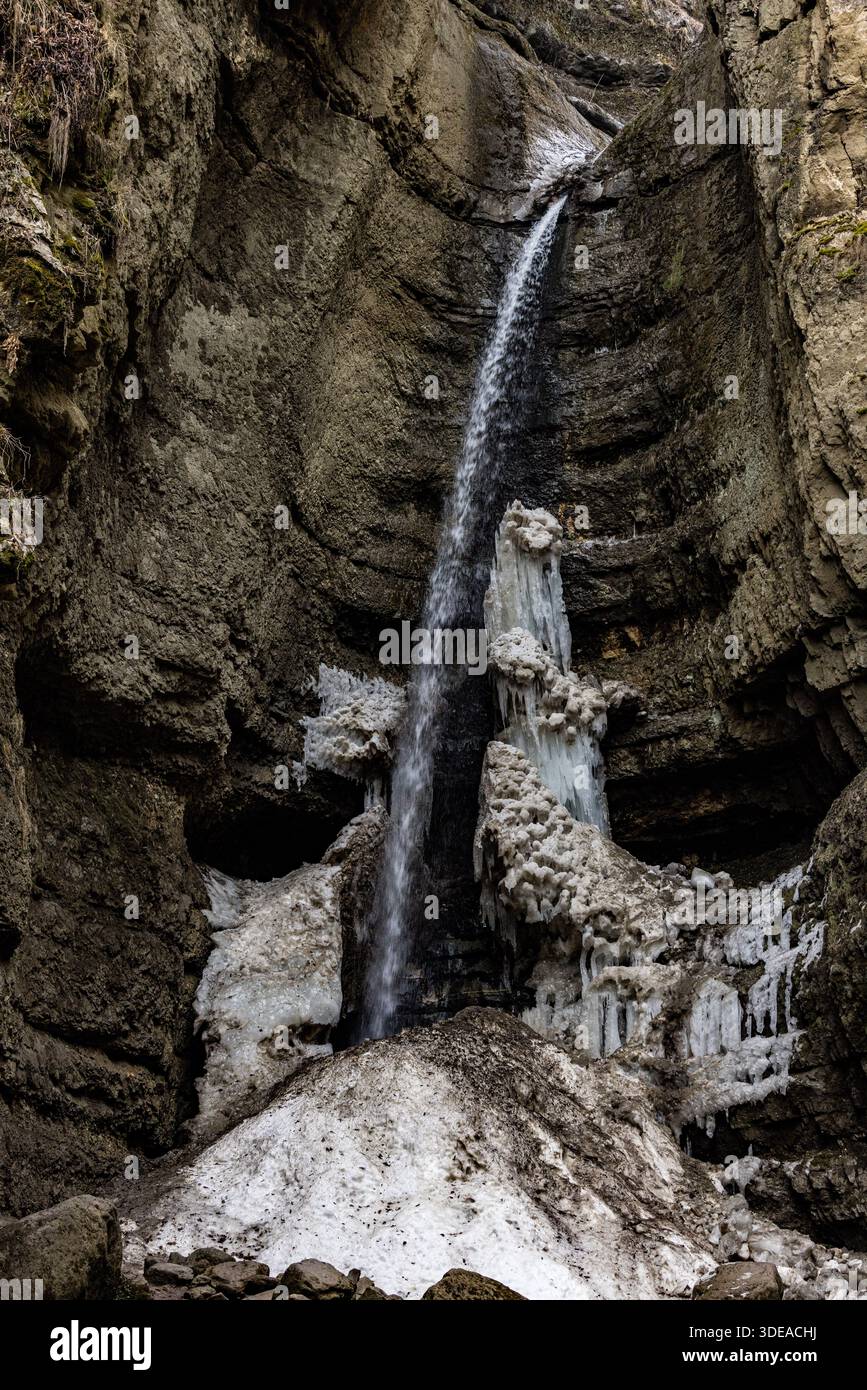 Piccola cascata parzialmente congelata all'interno di una stretta gola rocciosa. Paesaggio montano invernale con formazioni di ghiaccio e acqua che scorre. Foto Stock