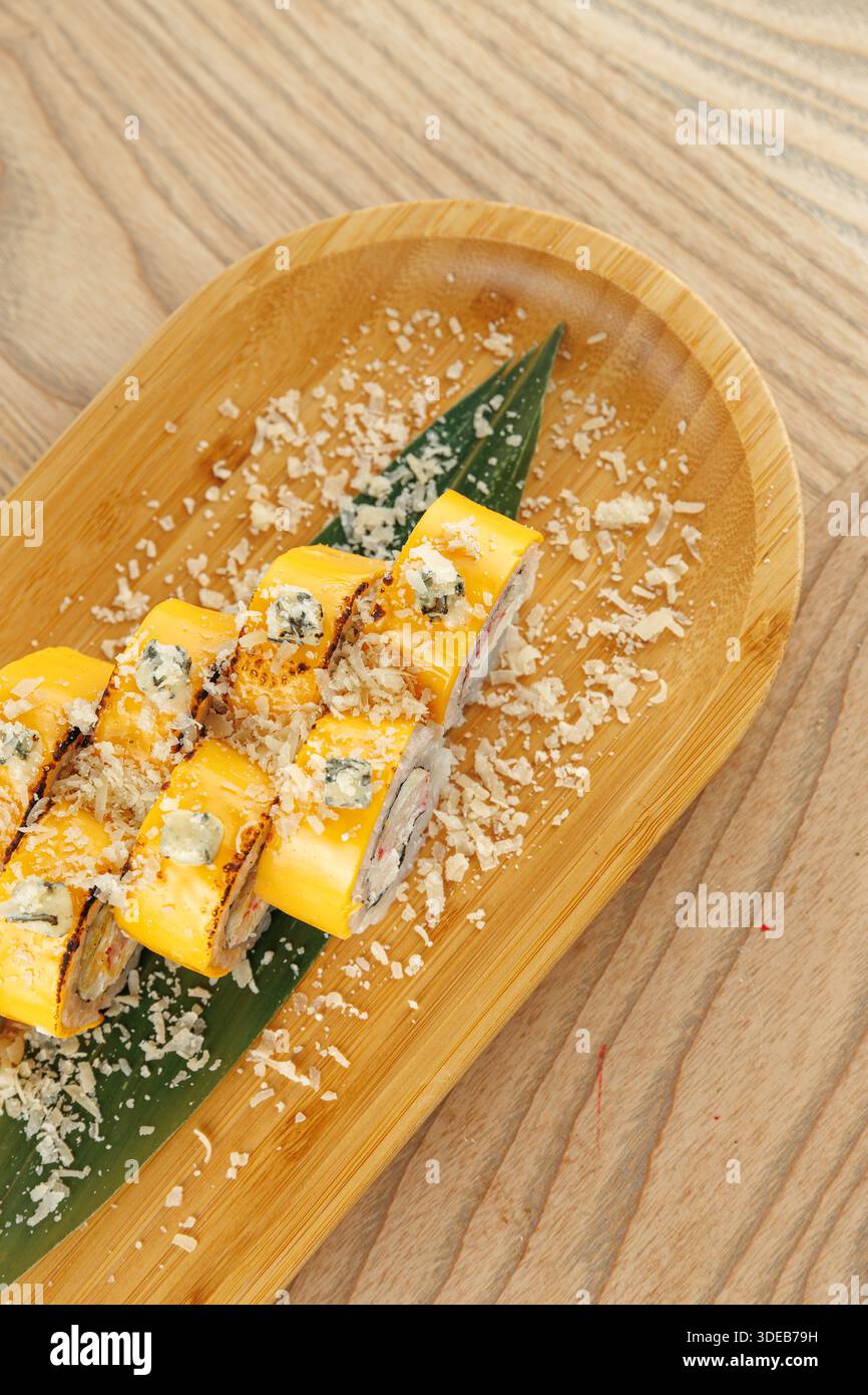 Gli involtini di sushi sono seduti su un piatto di bambù ricoperto di cocco grattugiato e posti su una foglia verde. L'ambientazione mostra un semplice tavolo di legno comune in giapponese di Foto Stock
