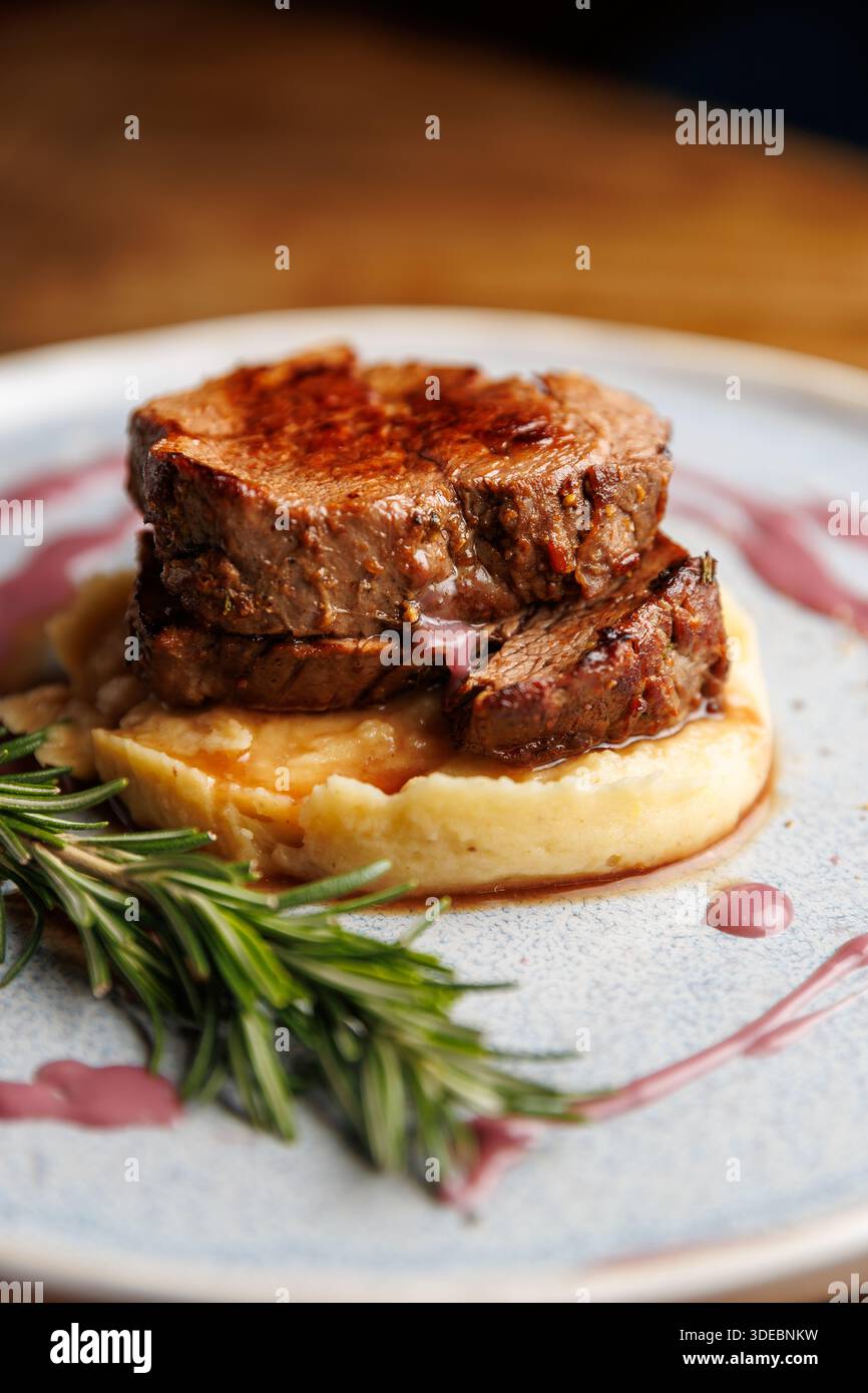 Il filetto di manzo cotto alla perfezione è impilato su purè di patate lisce, condito con salsa di vino rosso e guarnito con rosmarino fresco su ruggine Foto Stock