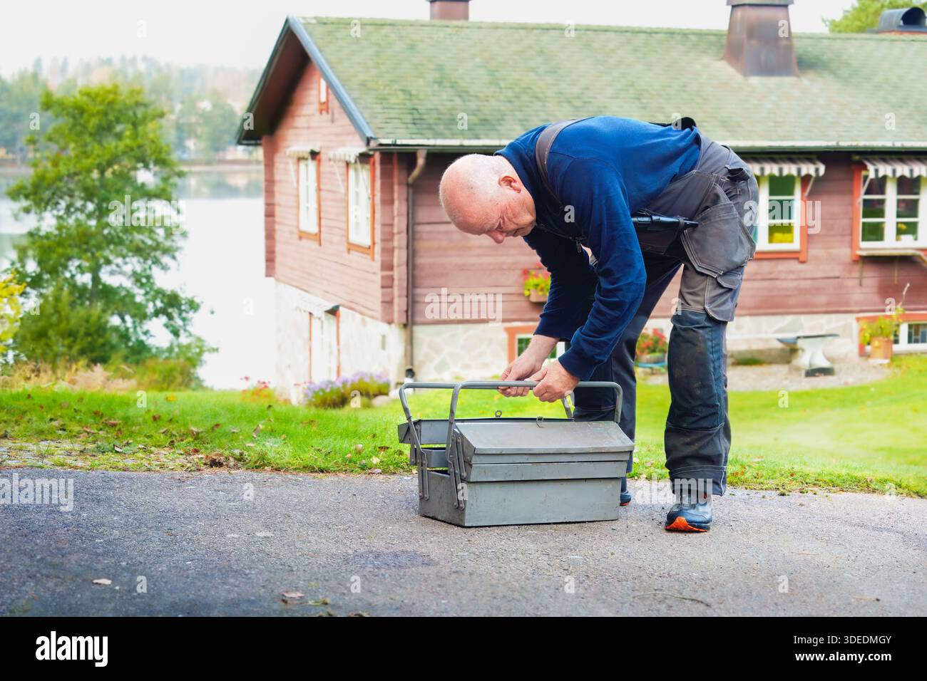 L'uomo anziano si concentra sull'organizzazione degli strumenti da una cassetta degli attrezzi all'aperto, con una casa di legno e verde sullo sfondo, evidenziando il miglioramento della casa Foto Stock
