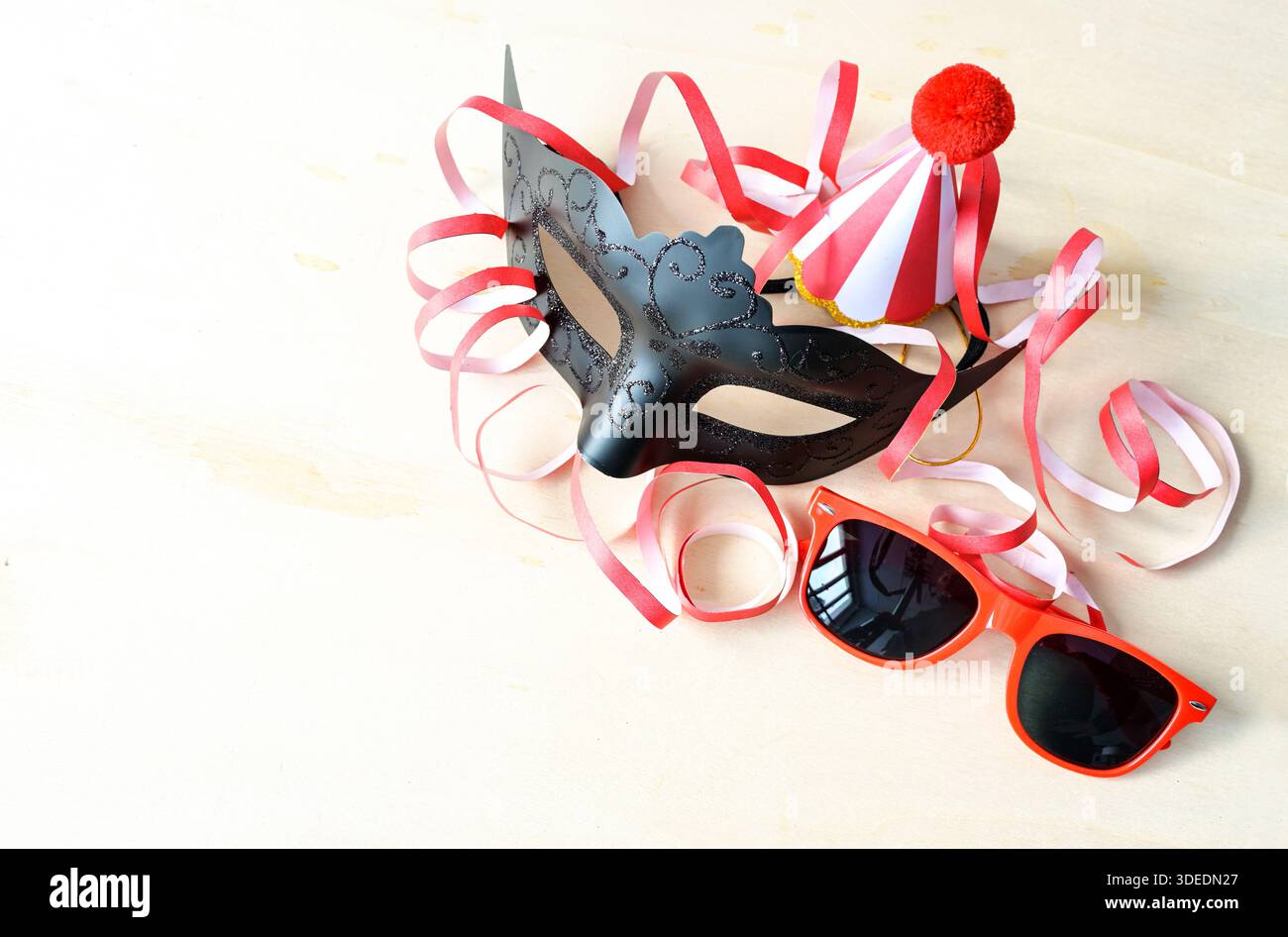 Accessori per feste di Carnevale, maschera nera, occhiali da sole rossi, cappello divertente e streamer su sfondo di legno chiaro, concept per eventi stagionali, invito Foto Stock