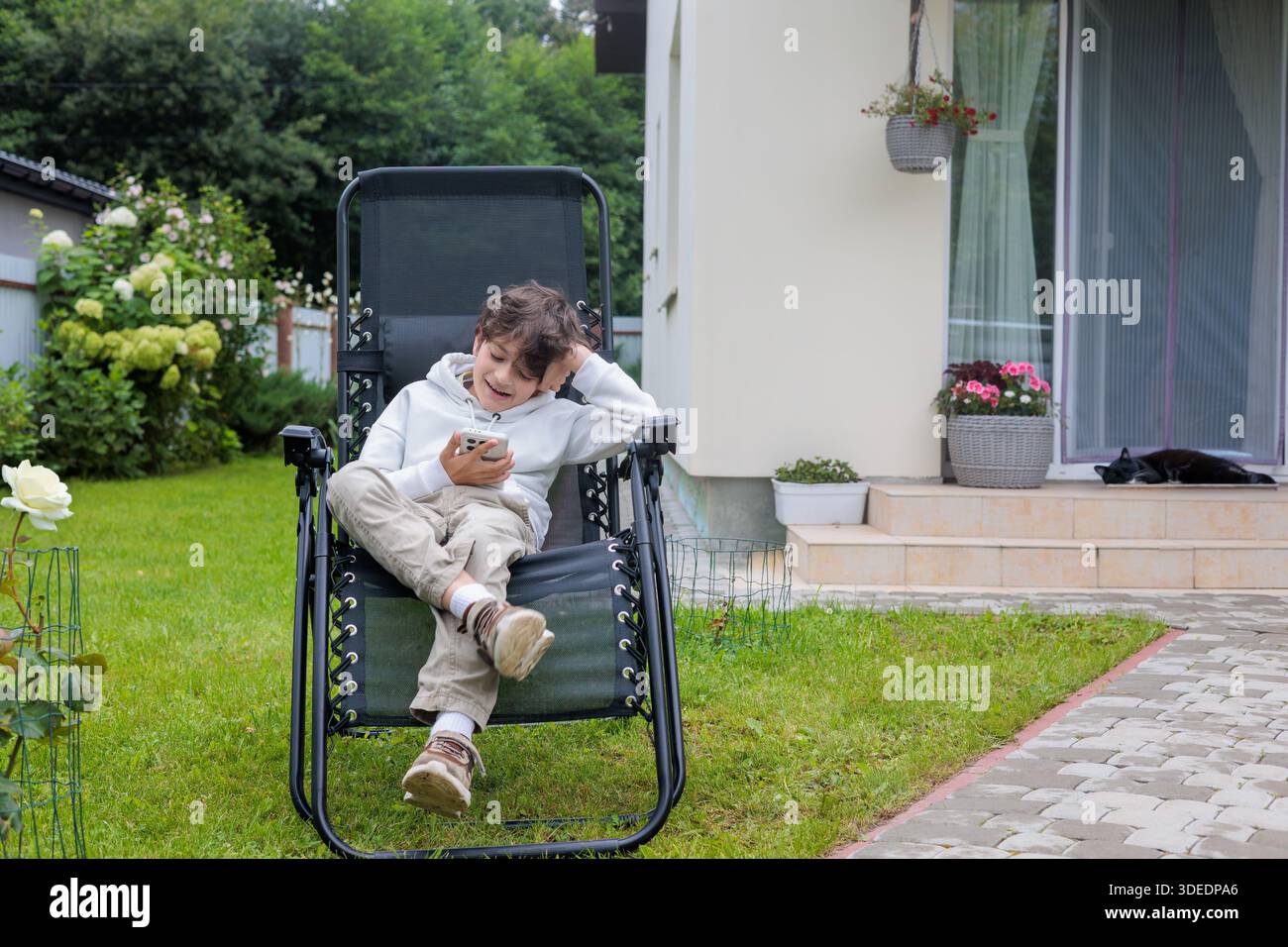 Bambino sorridente che si rilassa all'aperto sulla sedia da giardino utilizzando lo smartphone, lo stile di vita moderno dell'infanzia, il tempo libero digitale a casa, la tecnologia e lo schermo Foto Stock