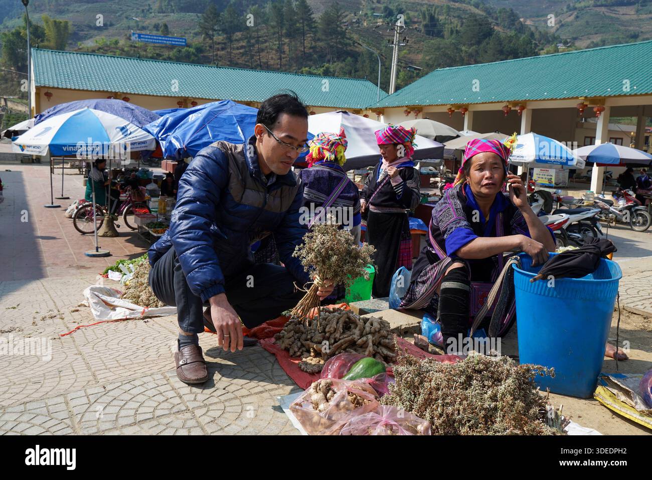Vivace mercato etnico in Vietnam: L'uomo esamina le erbe, la donna in abiti tradizionali parla al telefono tra prodotti freschi e commercio locale. Foto Stock