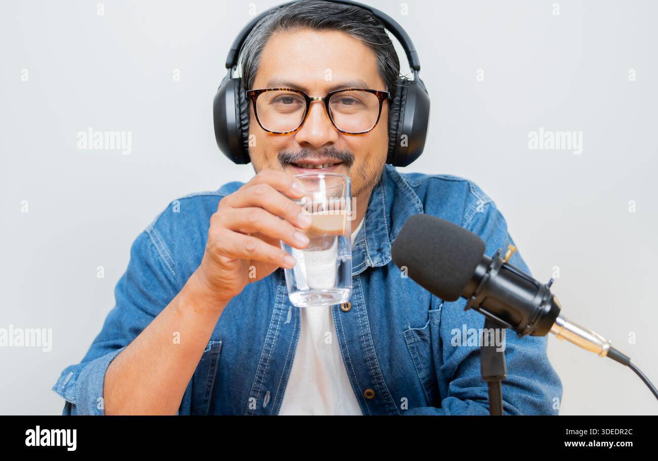 Bell'uomo barbuto che beve una tazza d'acqua mentre registra podcast con microfono professionale e guarda la fotocamera Foto Stock